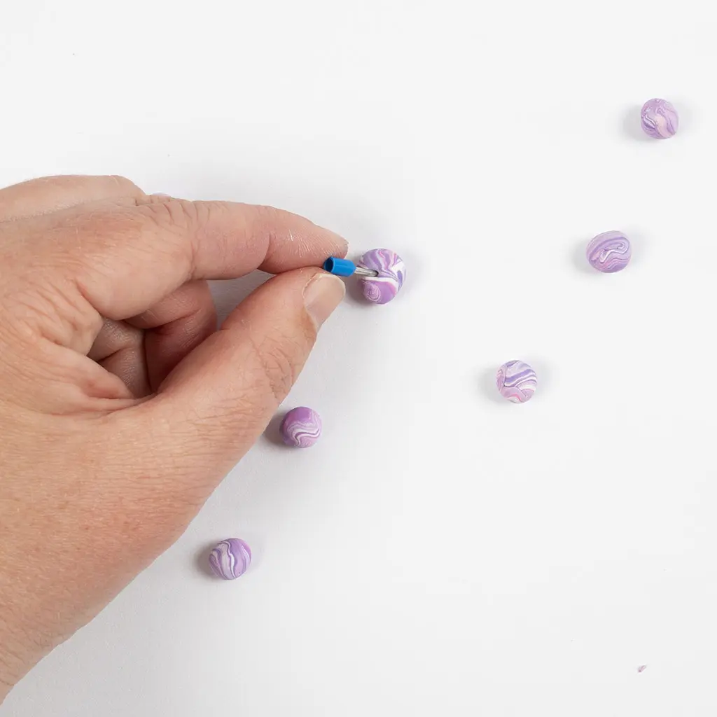 Een hand maakt met een blauw gereedschapje een gaatje in het midden van een gemarmerd paars kleibolletje, met andere afgewerkte kraaltjes eromheen.