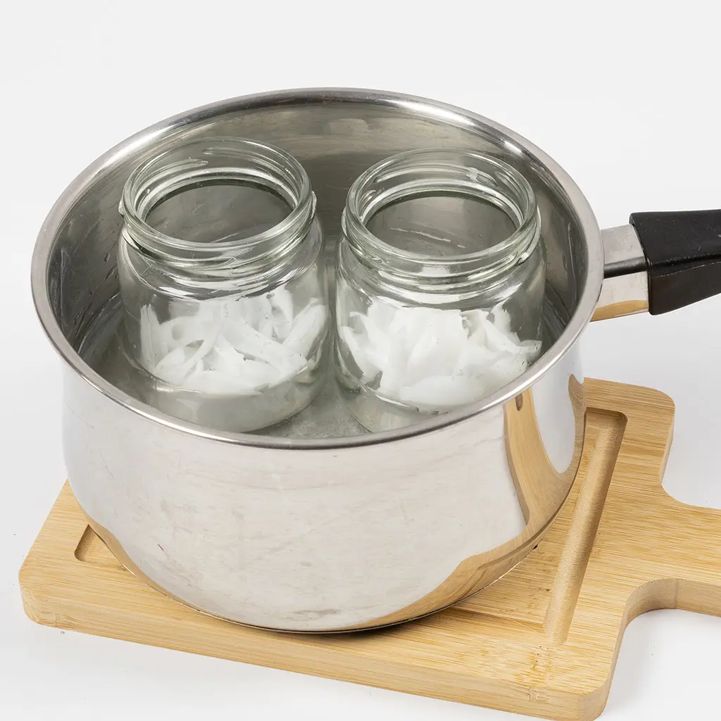 2 glazen potten met stukjes witte zeep staan in een pan met water op een houten rechthoekige bord.