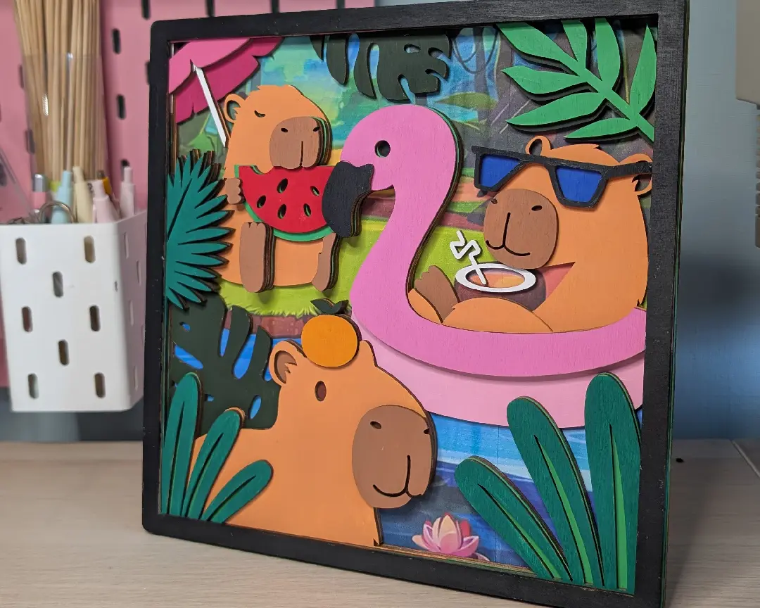 Volledig afgewerkt gelaagd houten capybara-kunstwerk in zwarte lijst, staand op een bureau met op de achtergrond penselenpotjes en een roze wand.