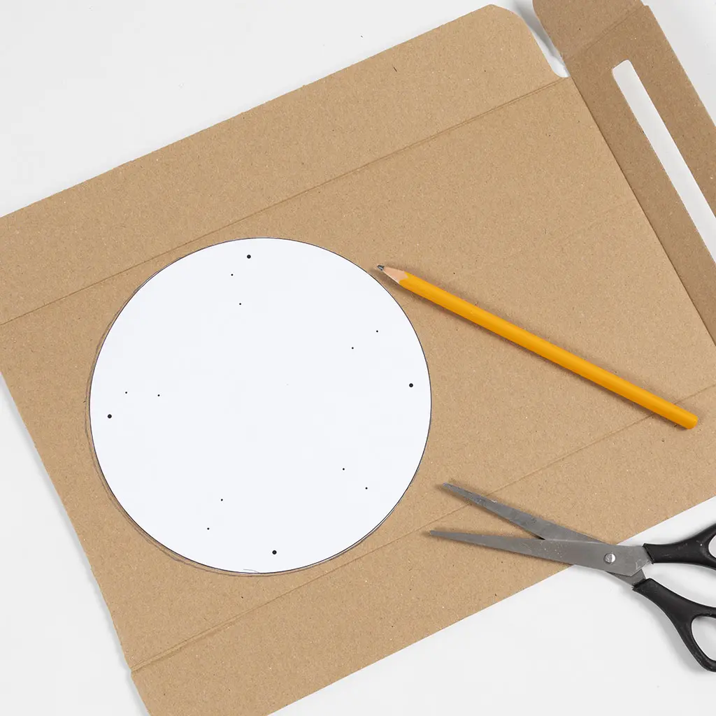Een wit rond sjabloon met stippen ligt op een stuk bruin karton. Er liggen een geel potlood en een hobbyschaar naast om de cirkel over te trekken en uit te knippen.