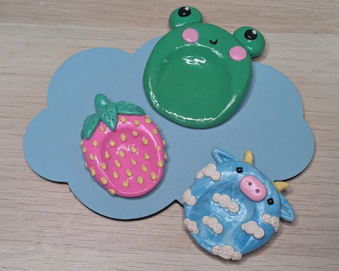 Drie voltooide, glanzend gelakte worry stones van polymeerklei. Een groene kikker, een roze aardbei en een blauw koetje met wolkjes, liggen gepresenteerd op een blauwe, wolkachtige onderzetter op een houten tafel.
