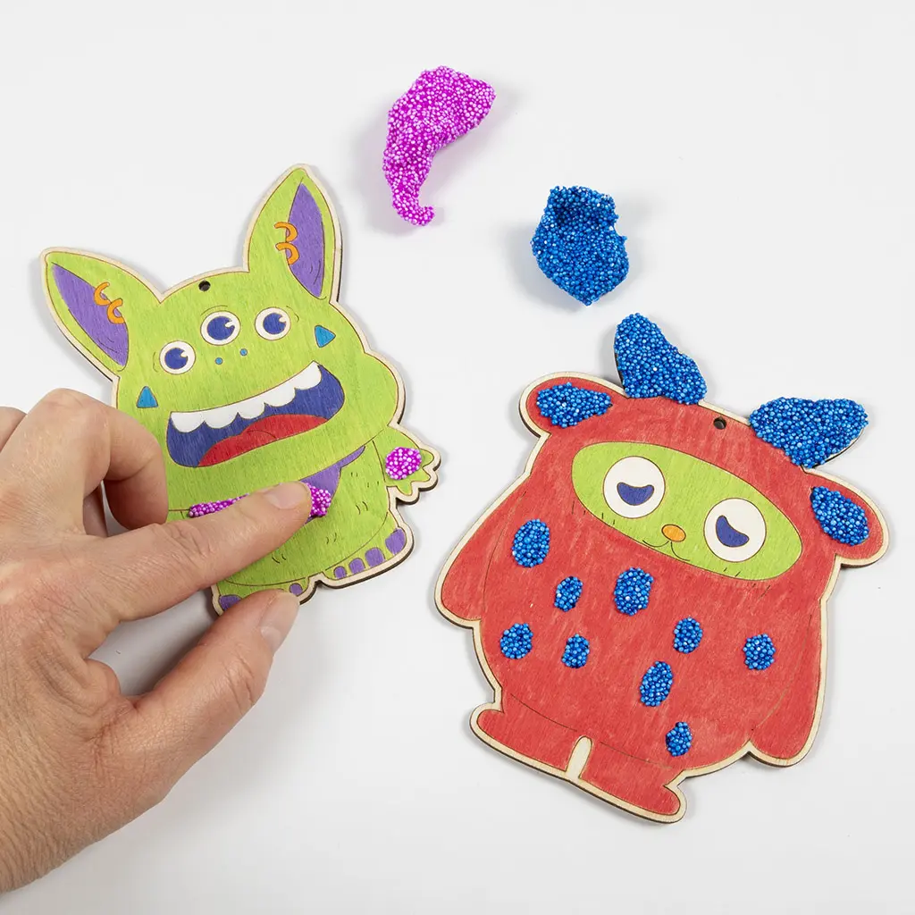 Een hand brengt roze Foam Clay aan op de mond en handen van het drieoogige groene monster. Ernaast ligt het rode monster, waarvan de hoorns en details op het lichaam zijn versierd met blauwe Foam Clay.