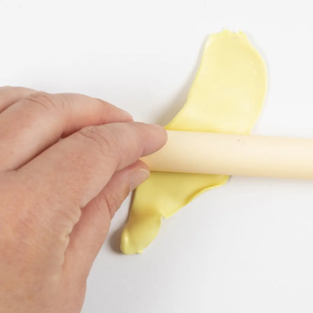 Een hand rolt de lichtgele klei plat met een crèmekleurige deegroller op een witte ondergrond.
