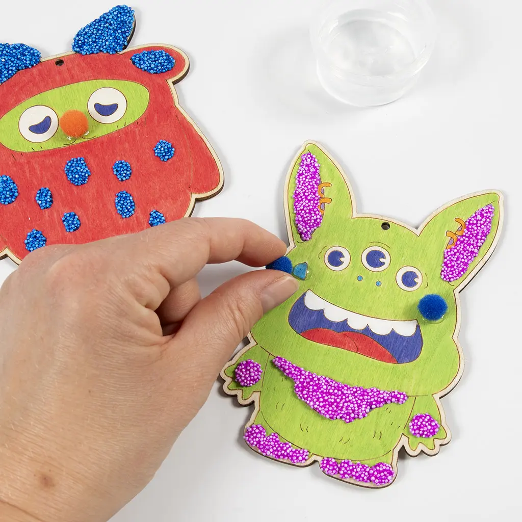 Een hand plakt een blauw pomponnetje als wang op het groene houten monster met vloeibare lijm. Op de achtergrond is bij het rode monster een oranje pompon als neus bevestigd.