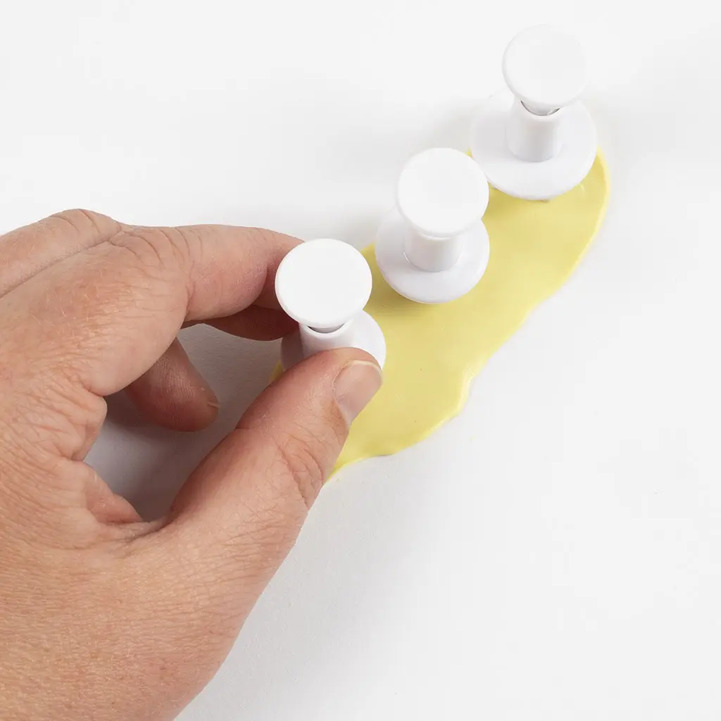 Een hand drukt drie witte stempeltjes/plungers in de platgerolde lichtgele klei om vormpjes uit te steken.