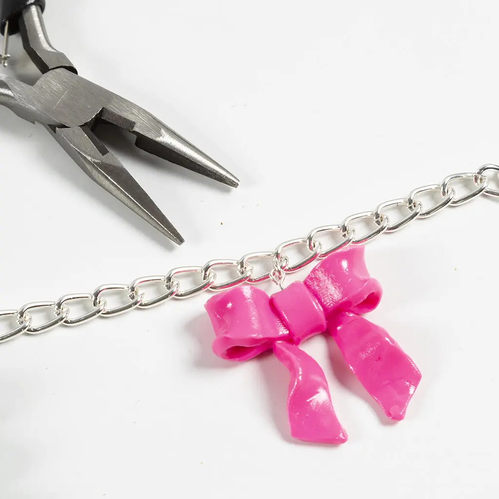 De glanzende roze kleistrik hangt als bedeltje aan een grove zilveren schakelketting. Linksboven ligt een tang op de witte achtergrond.