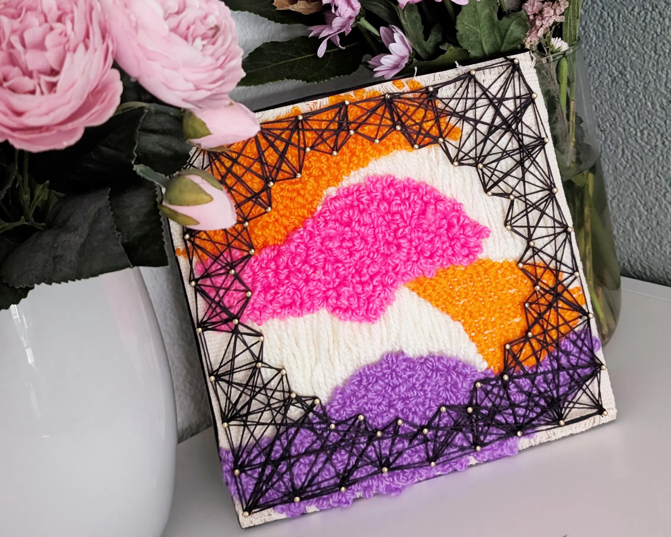 Afgewerkt combinatiekunstwerk van punch needle en string art met abstracte golvende vormen in oranje, roze en paars, omgeven door een geometrisch zwart garenpatroon, tentoongesteld naast roze bloemen
