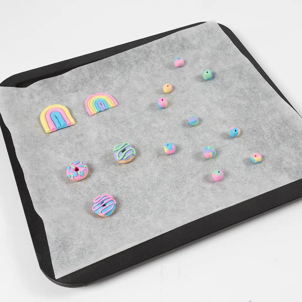 Een bakplaat met bakpapier waarop kleidonuts, regenboogjes en kleikralen liggen klaar om afgebakken te worden.