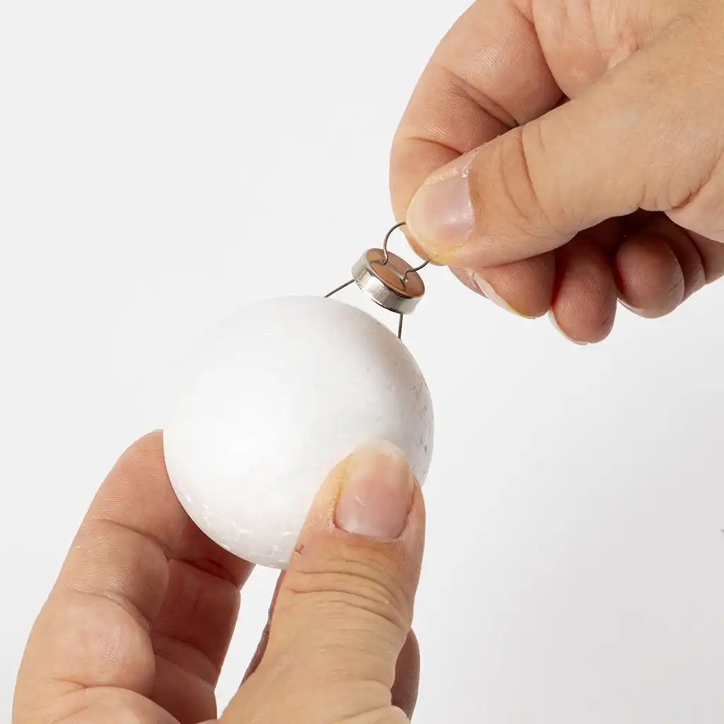Handschoenen duwen een metaalkleurig kroontje met een oogje bovenin een witte piepschuim bol.