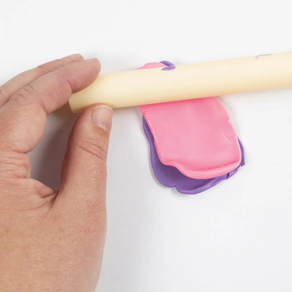 Een hand rolt een roze lapje klei plat over een paars lapje klei met een deegroller om twee lagen te combineren.
