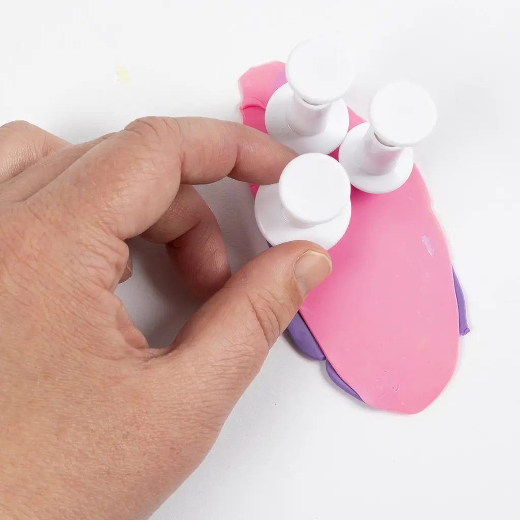 Een hand drukt drie witte uitstekers in de dubbele laag roze en paarse klei om meerkleurige vormpjes te maken.