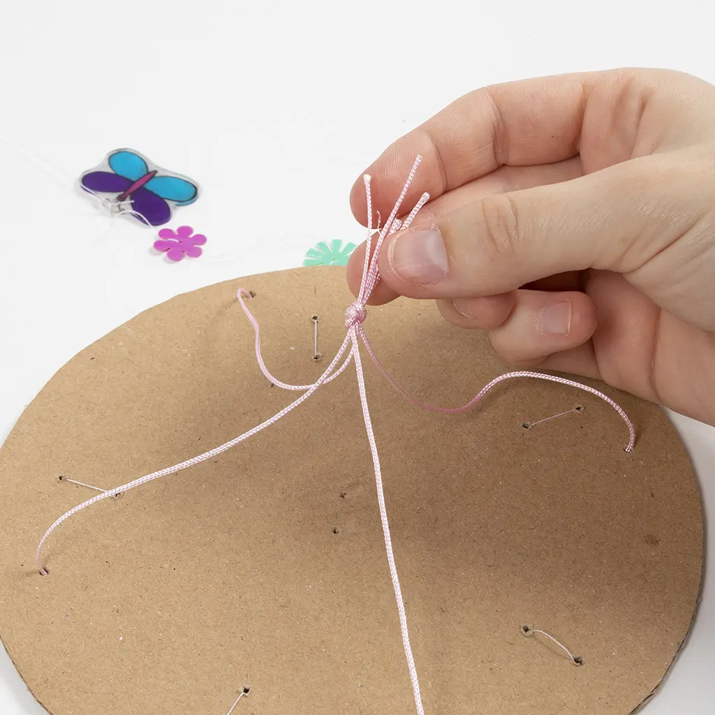 Een hand legt een knoop in de vier roze draden, zodat de mobiel stabiel en recht kan worden opgehangen.
