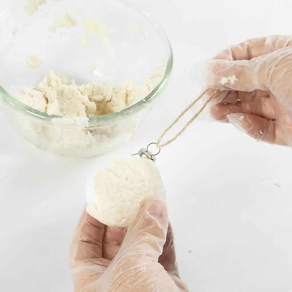 Een hand bedekt een witte piepschuim bol met een laagje natte papierpulp, met op de achtergrond een glazen mengkom met restanten pulp.
