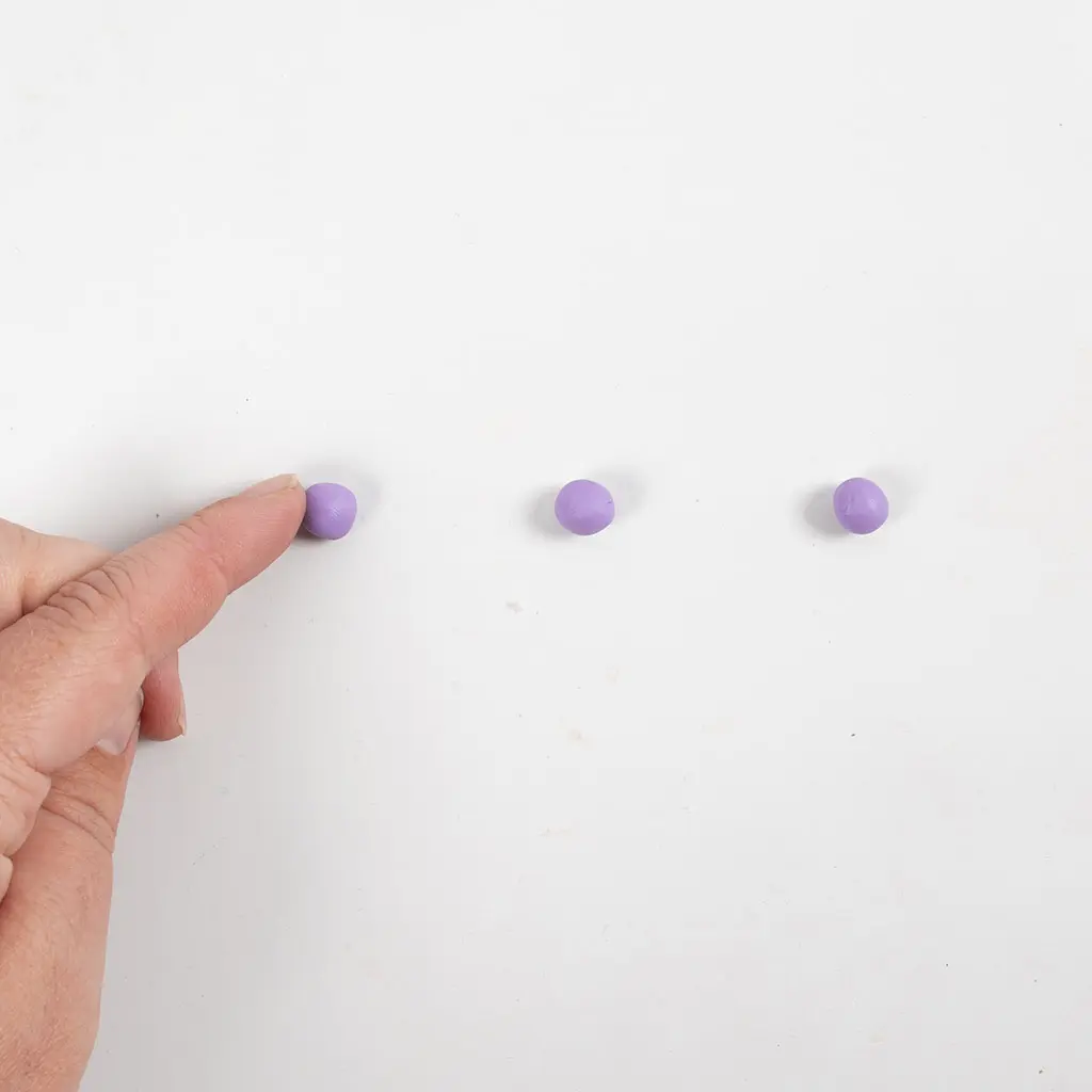 Een vinger wijst naar drie kleine, gelijke paarse kleibolletjes die op een rij op het werkoppervlak liggen.