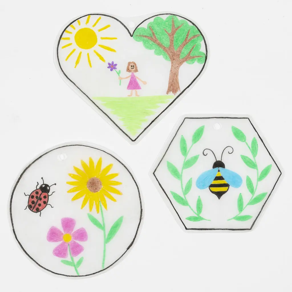 Drie platte, reeds gebakken krimpfolie-ontwerpen: een hartvorm met een meisje, de zon en een boom; een cirkel met een zonnebloem, roze bloem en lieveheersbeestje; en een zeshoek met een bij omringd door groene takjes.
