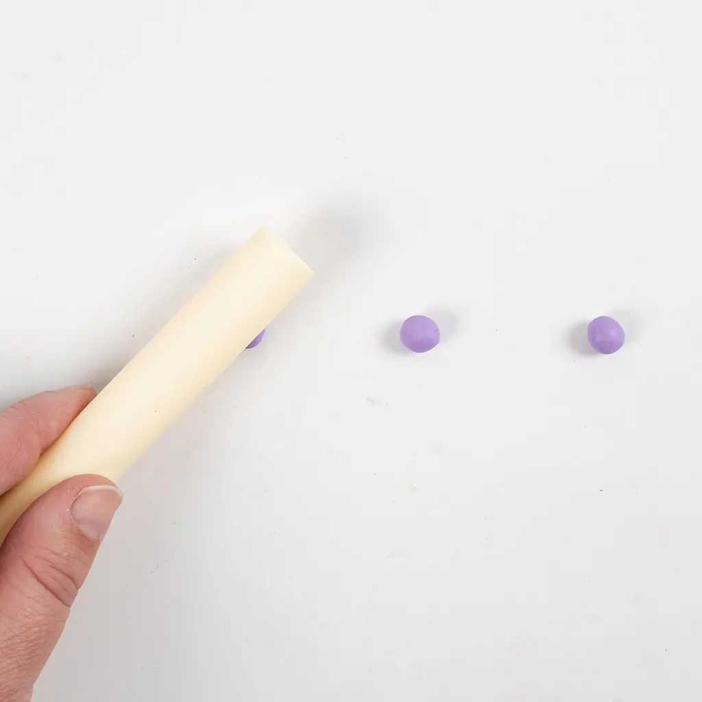Een hand rolt met een crèmekleurige roller het eerste paarse kleibolletje plat tot een schijfje.