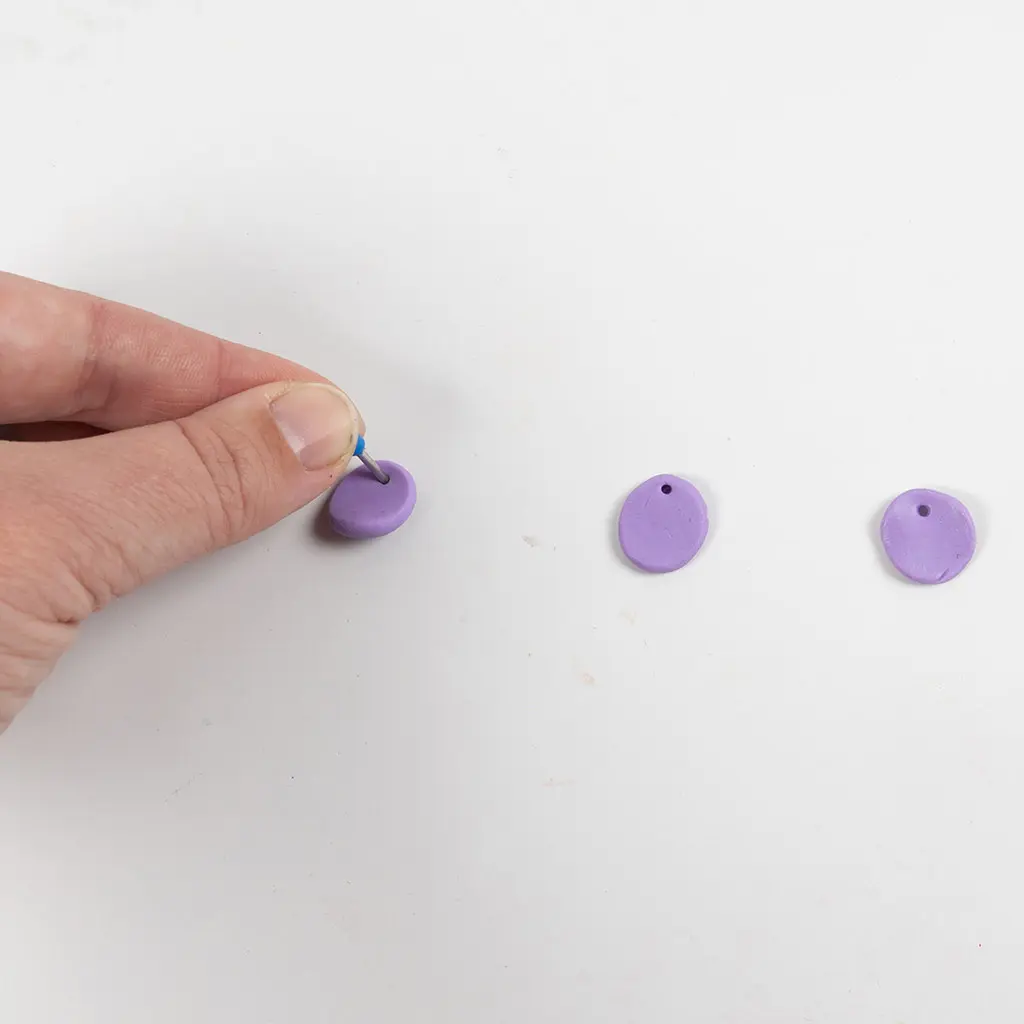 Een hand prikt met een blauwe pennenpunt een klein ophanggaatje aan de bovenkant van elk paars kleischijfje.