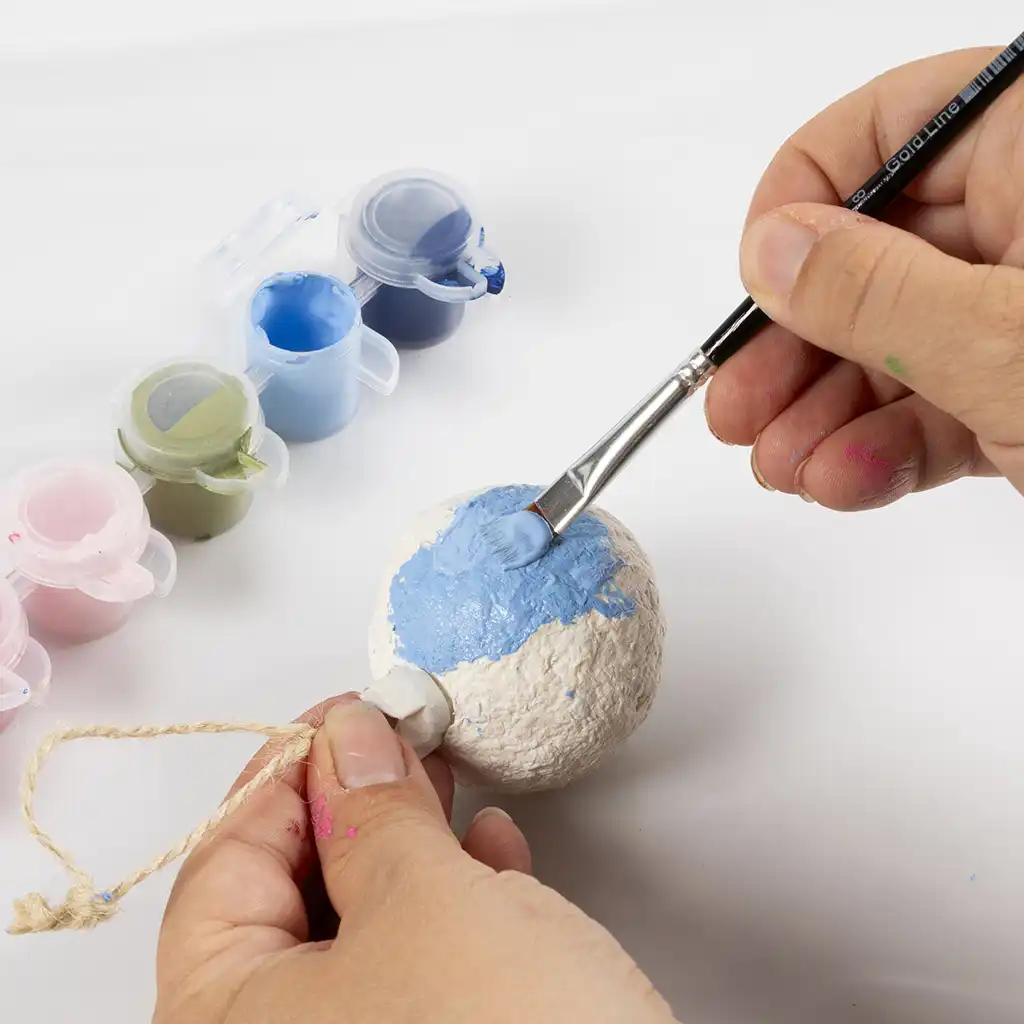 Een hand schildert de bal van papiermaché lichtblauw met een penseel, met op de achtergrond kleine verfpotjes.