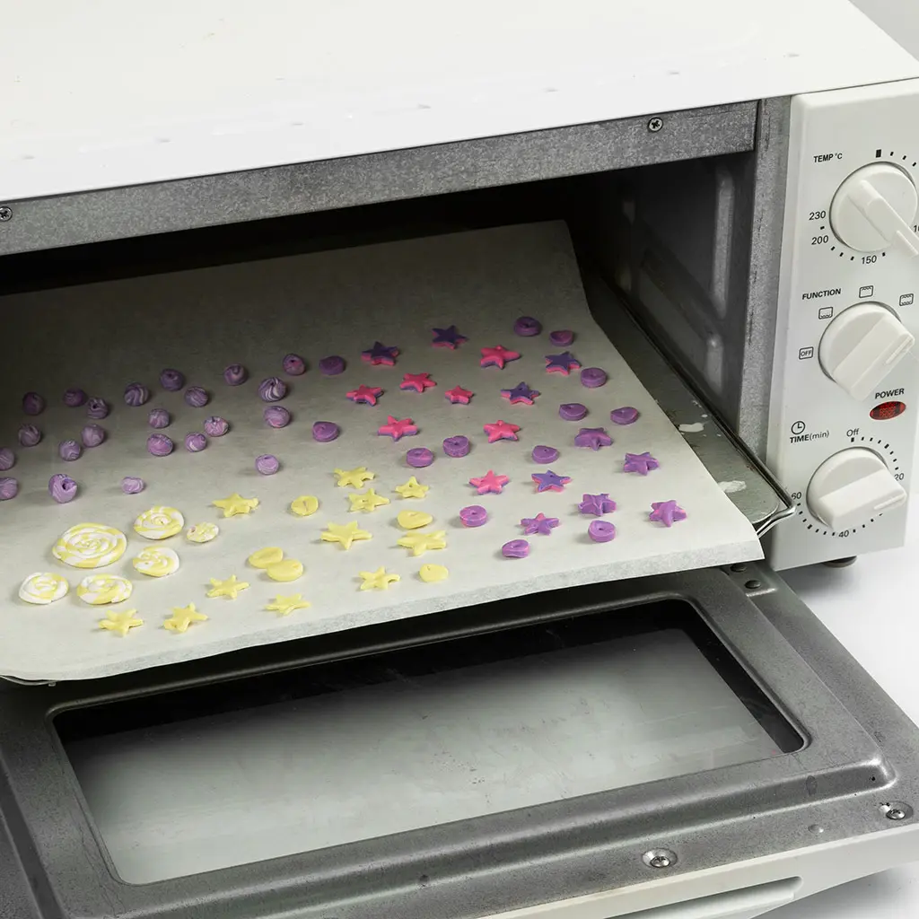 Een open mini-oven gevuld met een bakplaat vol gekleurde kleifiguren, sterren, lolly's en kraaltjes op bakpapier, klaar om afgebakken te worden.