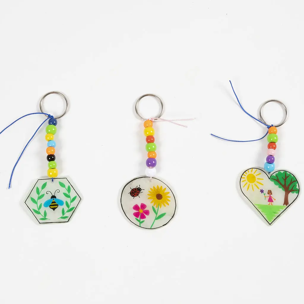 Drie voltooide sleutelhangers op een rij: de zeshoek met de bij, de cirkel met de bloemen en de hartvorm met het meisje. Alle drie zijn ze afgewerkt met een reeks kleurrijke kralen en een sleutelring.