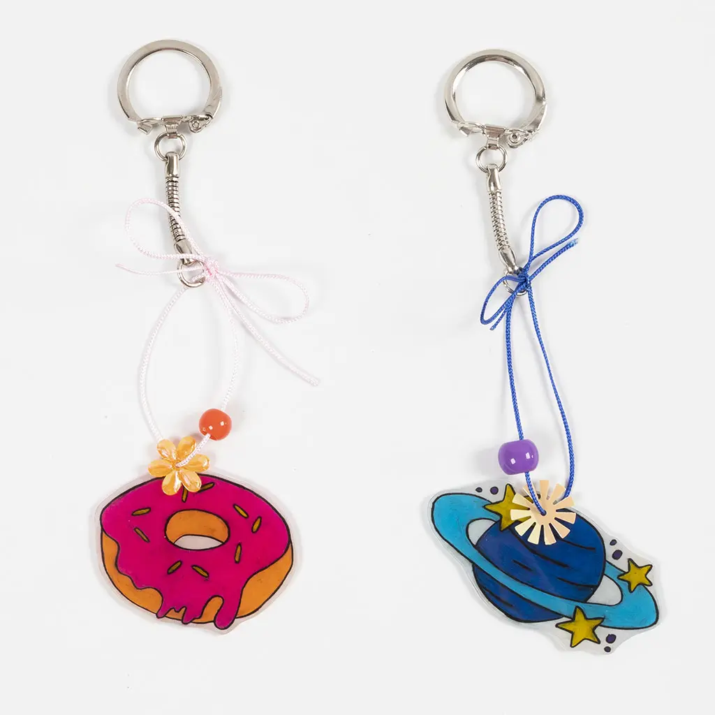 Twee voltooide sleutelhangers met ketting naast elkaar: links een felroze donut met een oranje kraal en bloemetje aan een roze koord, en rechts de planeet Saturnus aan een blauw koord. Beide koordjes zijn met een sierlijk strikje afgewerkt.
