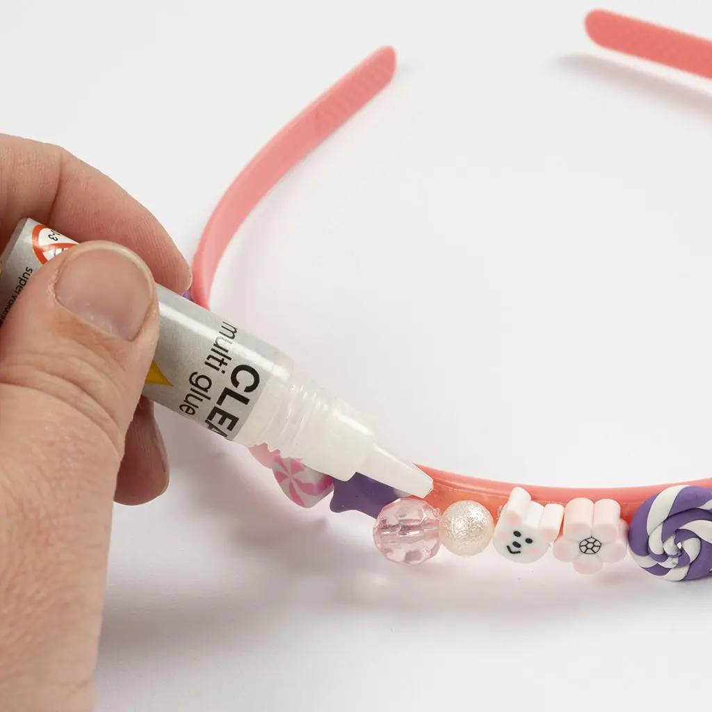 Een hand brengt extra transparante knutsellijm aan uit een tube om de kleidecoraties en kralen stevig op de haarband te fixeren.