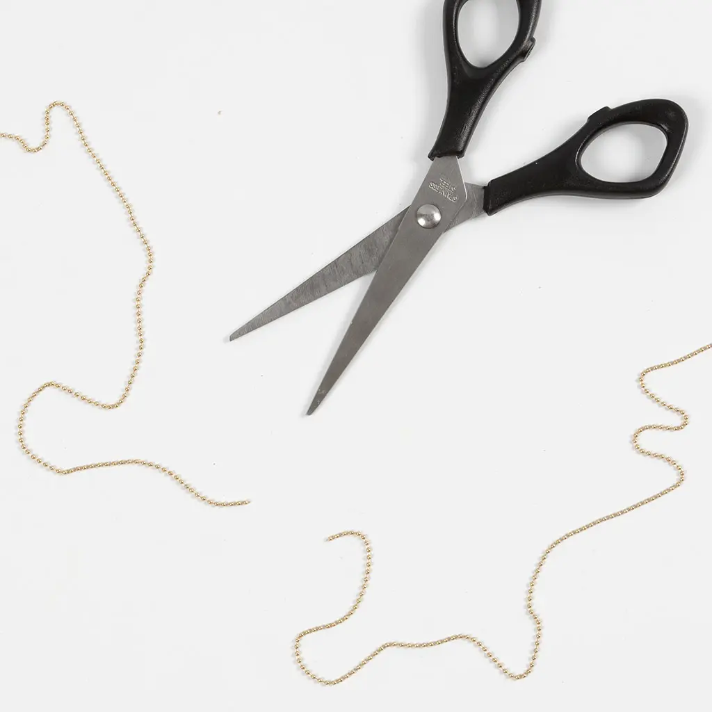 Een metalen hobbyschaar ligt op een witte ondergrond te midden van enkele op maat geknipte stukken goudkleurige bolletjesketting.