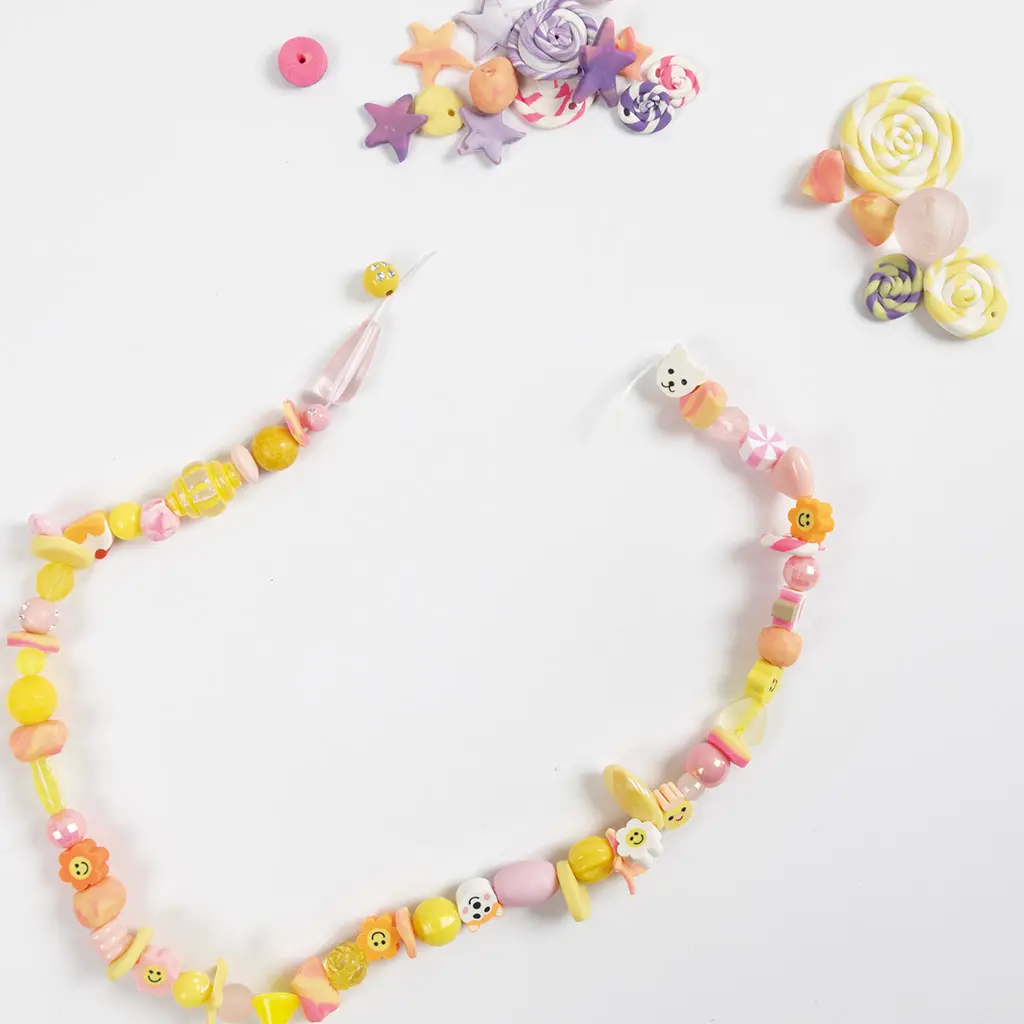 Een kleurrijke geregen kralenketting met afgebakken kleikraaltjes, sterren en vrolijke figuurtjes op een witte achtergrond, omringd door losse kleielementen.
