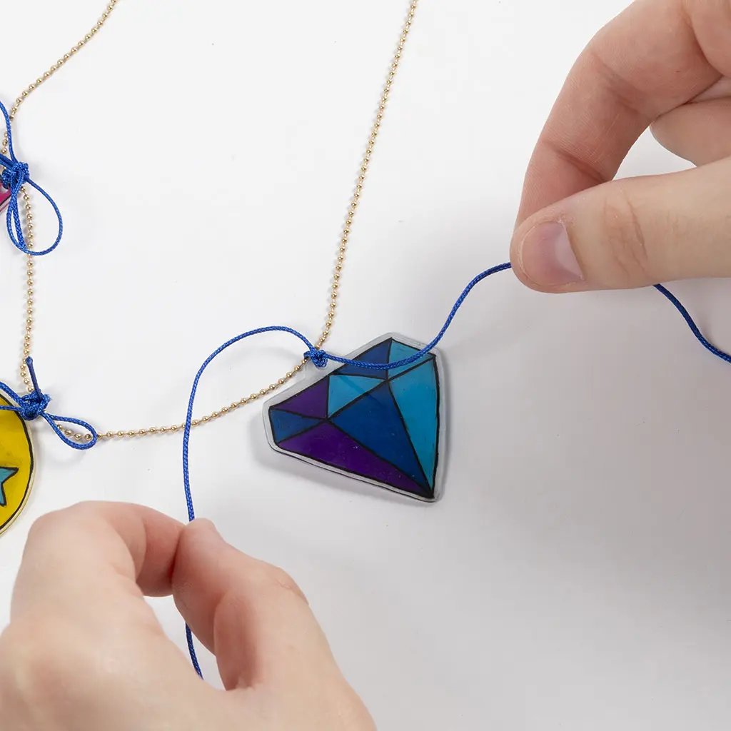 Twee handen knopen een blauw draadje rond een goudkleurige bolletjesketting om een diamantvormige hanger van krimpfolie (in blauw- en paarstinten) te bevestigen als sieraad.
