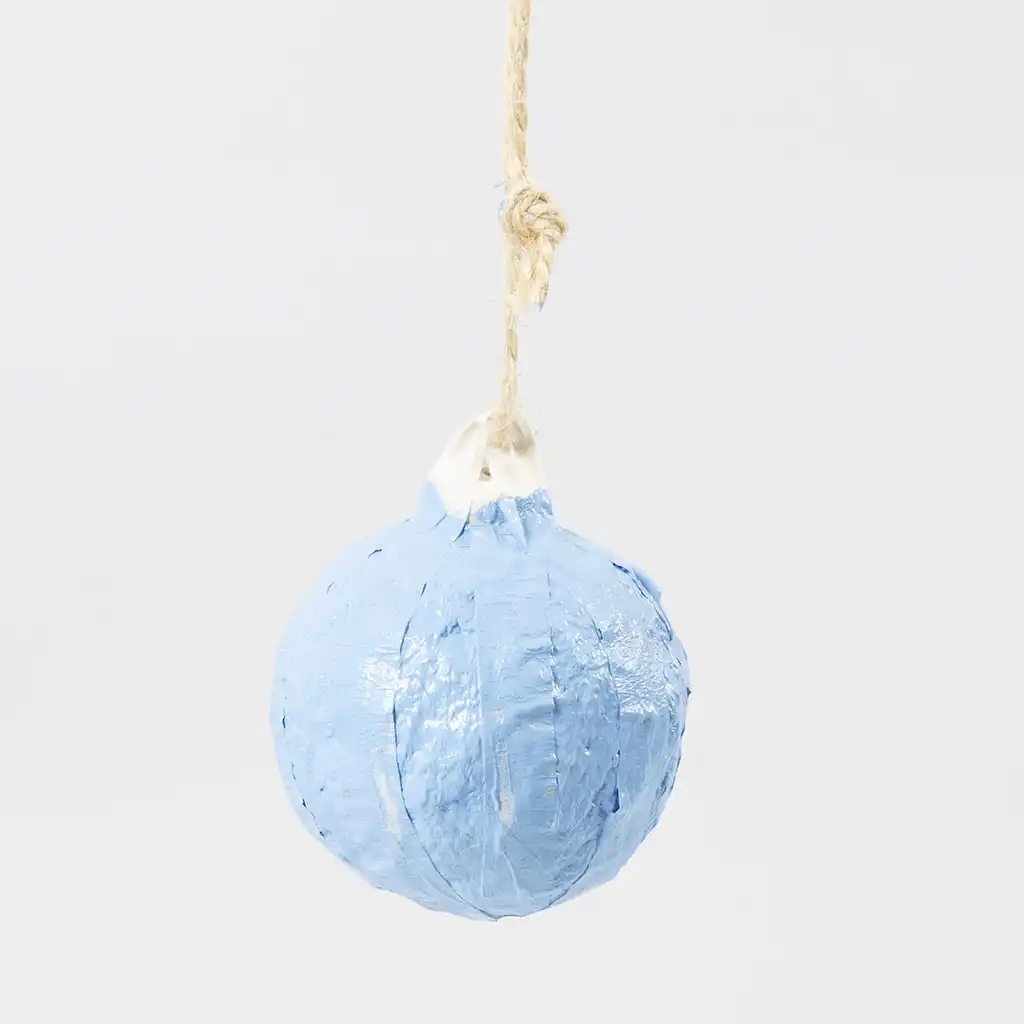 De met tape afgeplakte lichtblauwe kerstbal van papiermaché hangt te drogen aan het jute touwtje op een witte achtergrond.