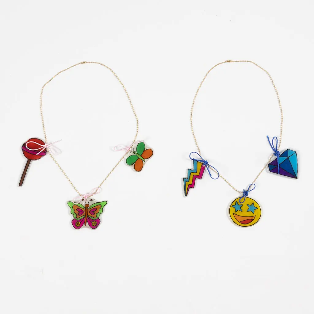 Twee voltooide kettingen met krimpfoliehangers naast elkaar. De linker ketting heeft een lolly, een grote vlinder en een kleine vlinder met roze strikjes. De rechter ketting heeft een bliksemschicht, een smiley met sterrenogen en een diamant met blauwe strikjes.
