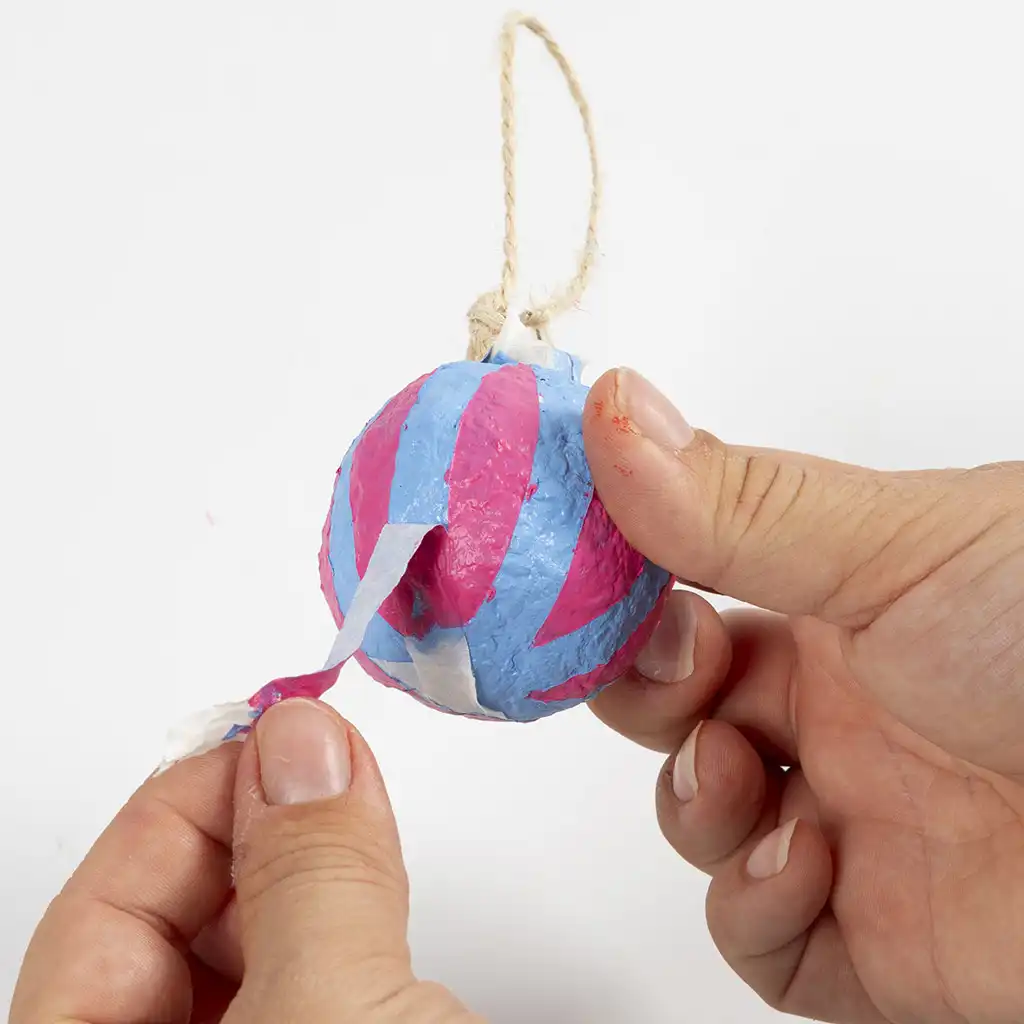 Twee handen trekken voorzichtig de stroken schilderstape van de kerstbal af, waardoor een blauw met roze gestreept patroon tevoorschijn komt.