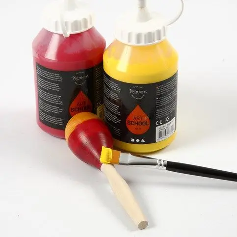 Een houten maraca wordt met een penseel geschilderd in de kleuren rood en geel, met twee grote flessen acrylverf op de achtergrond.