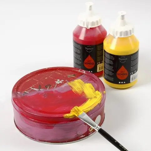 Een ronde blikken doos wordt met een penseel geschilderd in rood en geel, met twee grote flessen acrylverf op de achtergrond.
