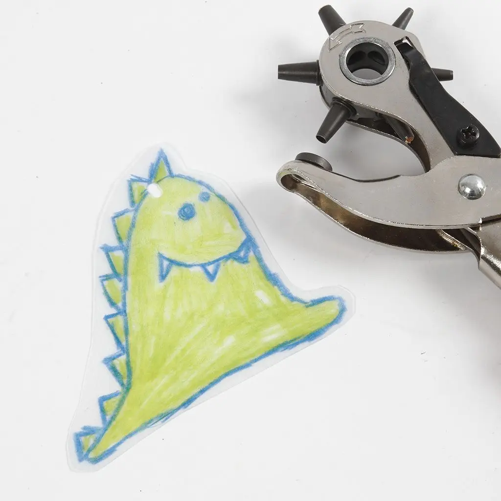 Een uitgeknipte, groen ingekleurde dinosaurus van krimpfolie met een klein gaatje aan de bovenkant. Rechtsboven ligt een metalen revolvertang (gaatjestang) op een witte achtergrond.
