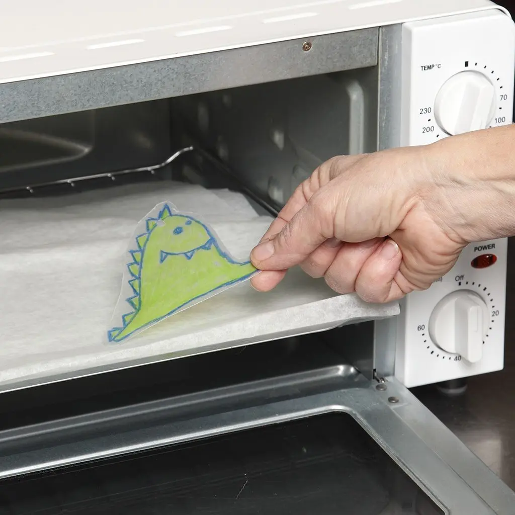 Een hand schuift een vel bakpapier met daarop de uitgeknipte dinosaurus van krimpfolie in een kleine, witte elektrische mini-oven om te krimpen.