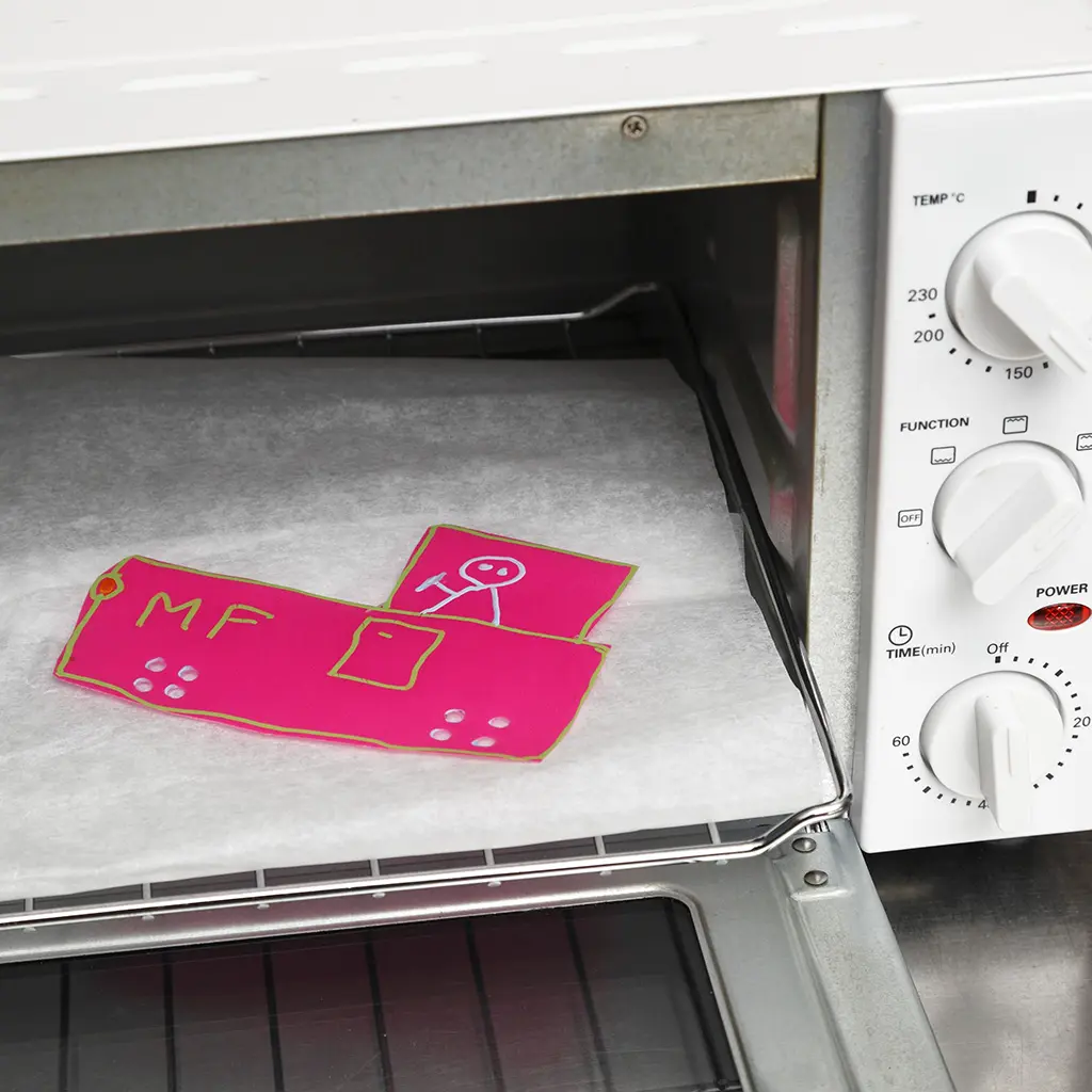 De roze krimpfolie-vrachtwagen ligt op een vel wit bakpapier op het ovenrooster, klaar om gebakken en gekrompen te worden in een witte mini-oven.