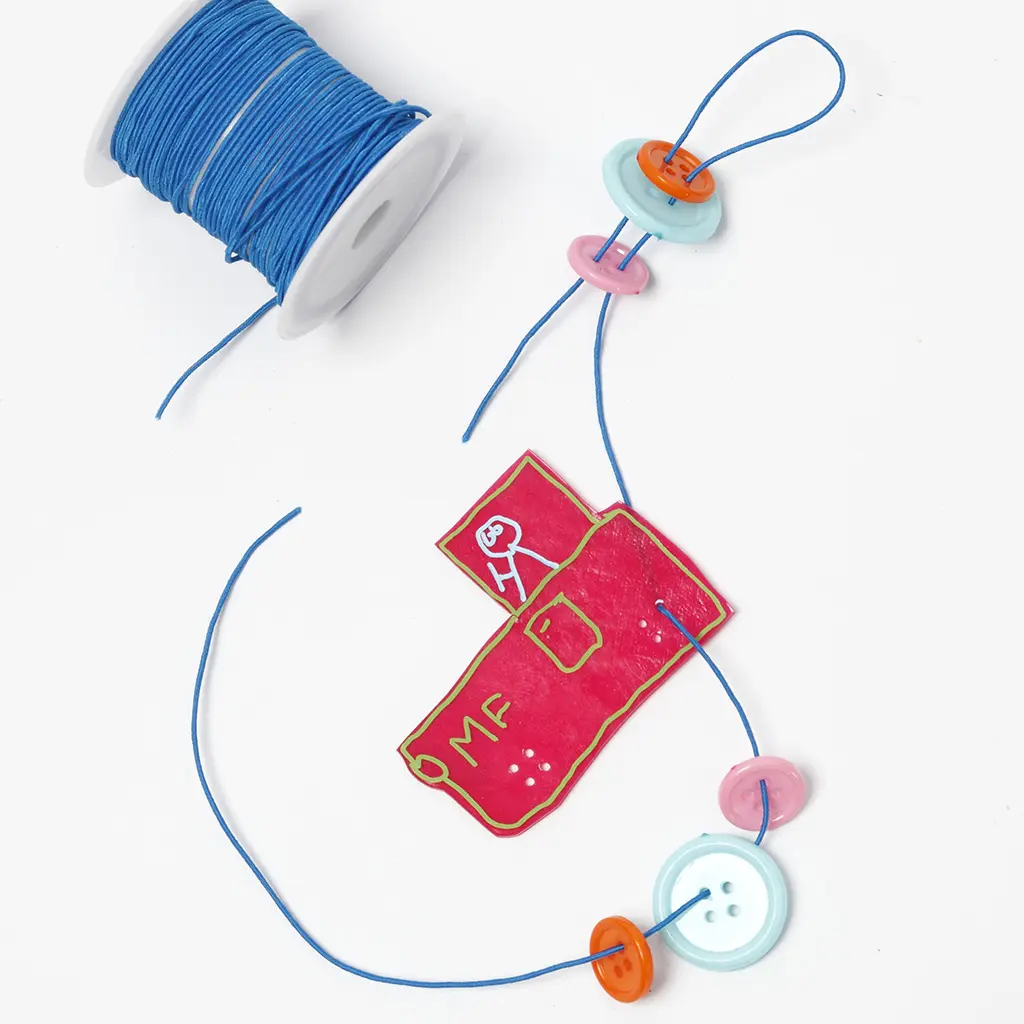 Een blauw koord wordt door de gaatjes van de inmiddels gekrompen, stevige roze vrachtwagen geregen. Ernaast liggen verschillende plastic knopen en een witte klos met blauw elastisch draad.