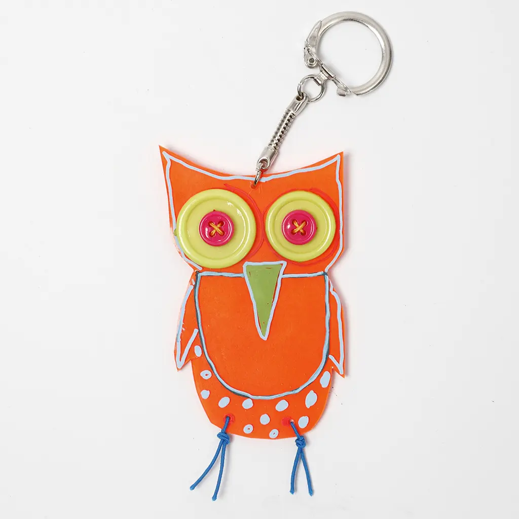 Een voltooide sleutelhanger van een uil, gemaakt van feloranje krimpfolie. De uil heeft grote ogen gemaakt van gele en roze knopen, blauwe details en pootjes van blauw draad.