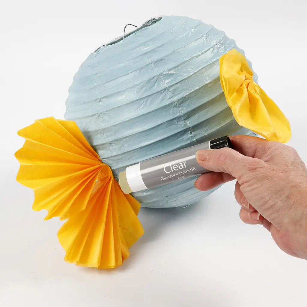 De gele geplooide staartvin wordt met een lijmstift aan de achterzijde van de blauwe lampion geplakt.