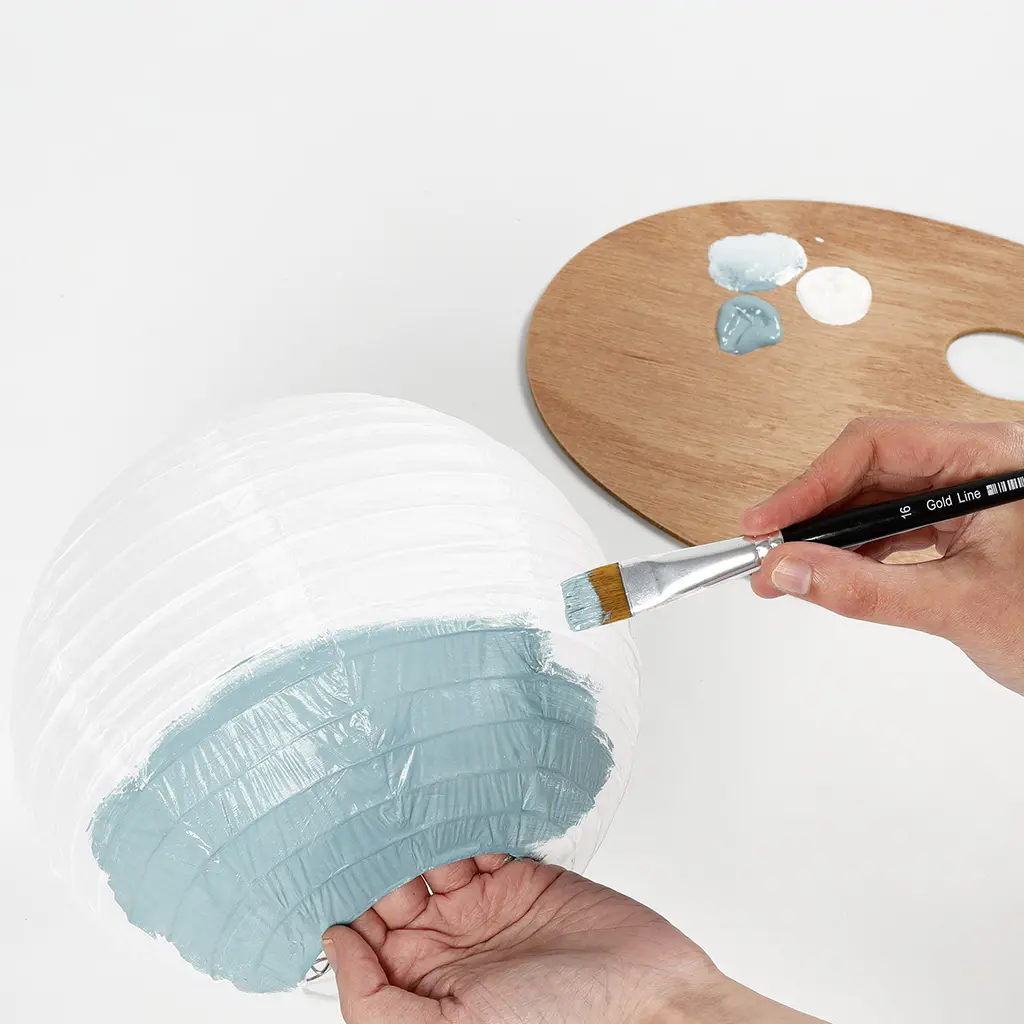 Een ronde witte papieren lampion wordt aan de onderkant deels lichtblauw geschilderd met een platte penseel.