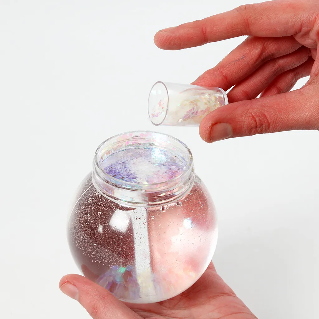 Een hand die fijne glitter- en sneeuwvlokjes uit een klein maaatbekertje in een transparante bol vol water giet.
