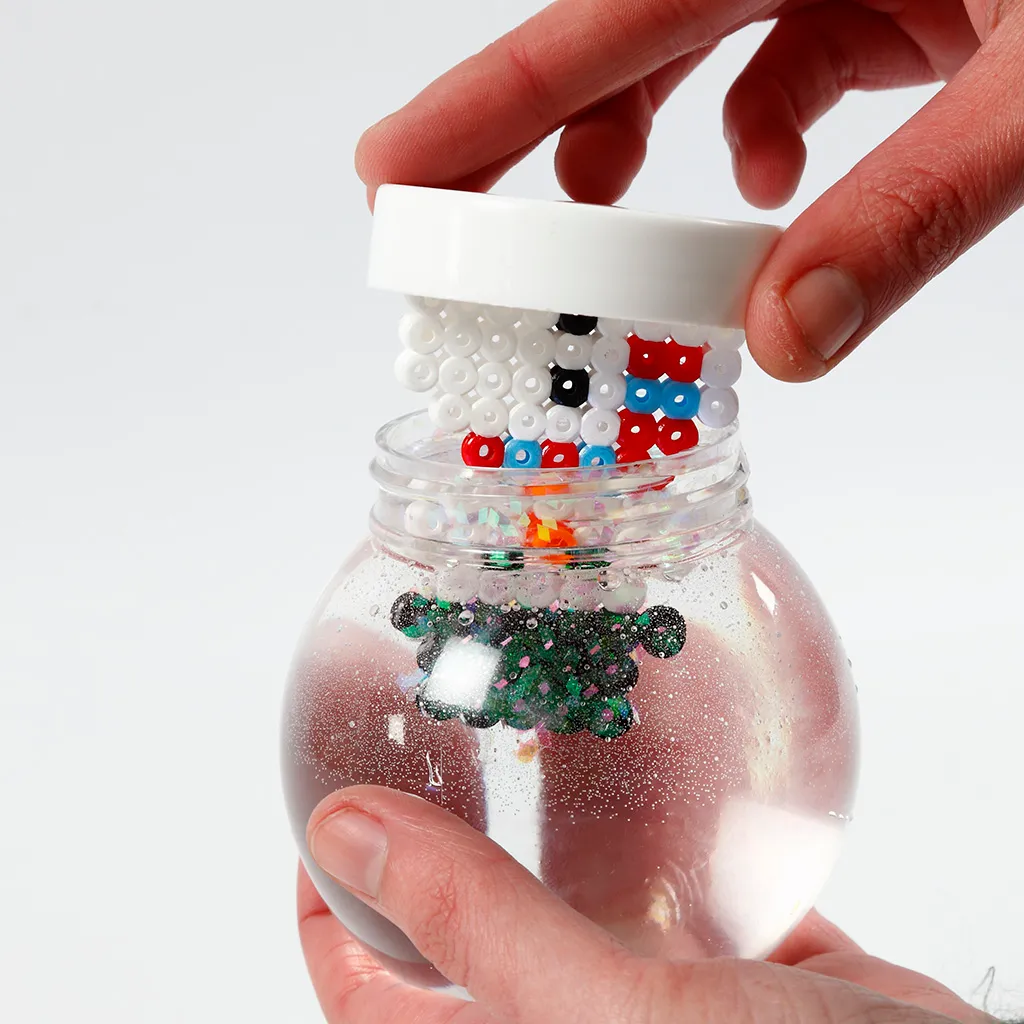 Een hand die de witte deksel met daaraan vastgemaakte strijkkraalfiguren omgekeerd in de met water en glitter gevulde bol plaatst.