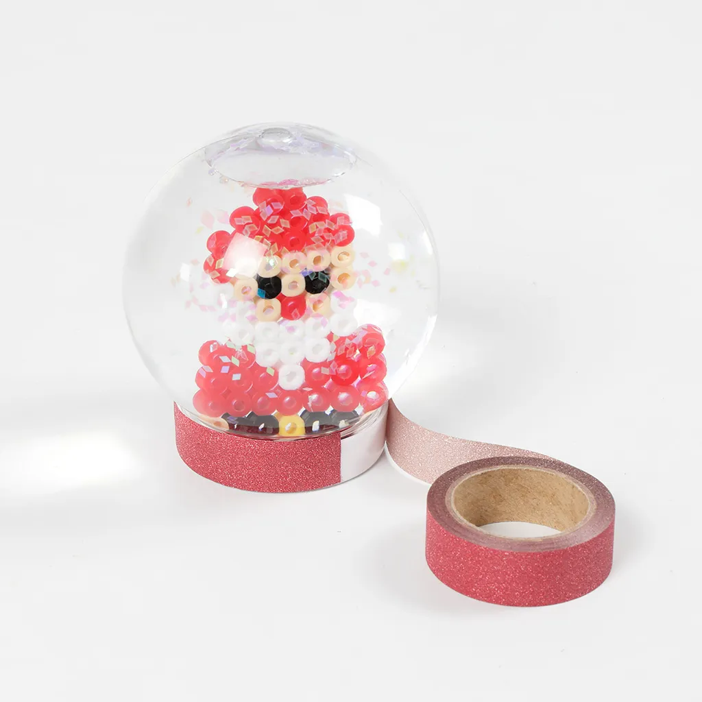 Een afgewerkte sneeuwbol met een kerstman van strijkkraaltjes, waarbij de onderkant wordt gedecoreerd met roze glitter-washi-tape.