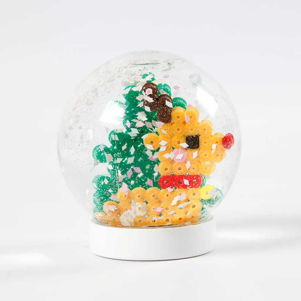 Een voltooide sneeuwbol op een witte voet met een rendier en een kerstboom van strijkkraaltjes in de sneeuw.