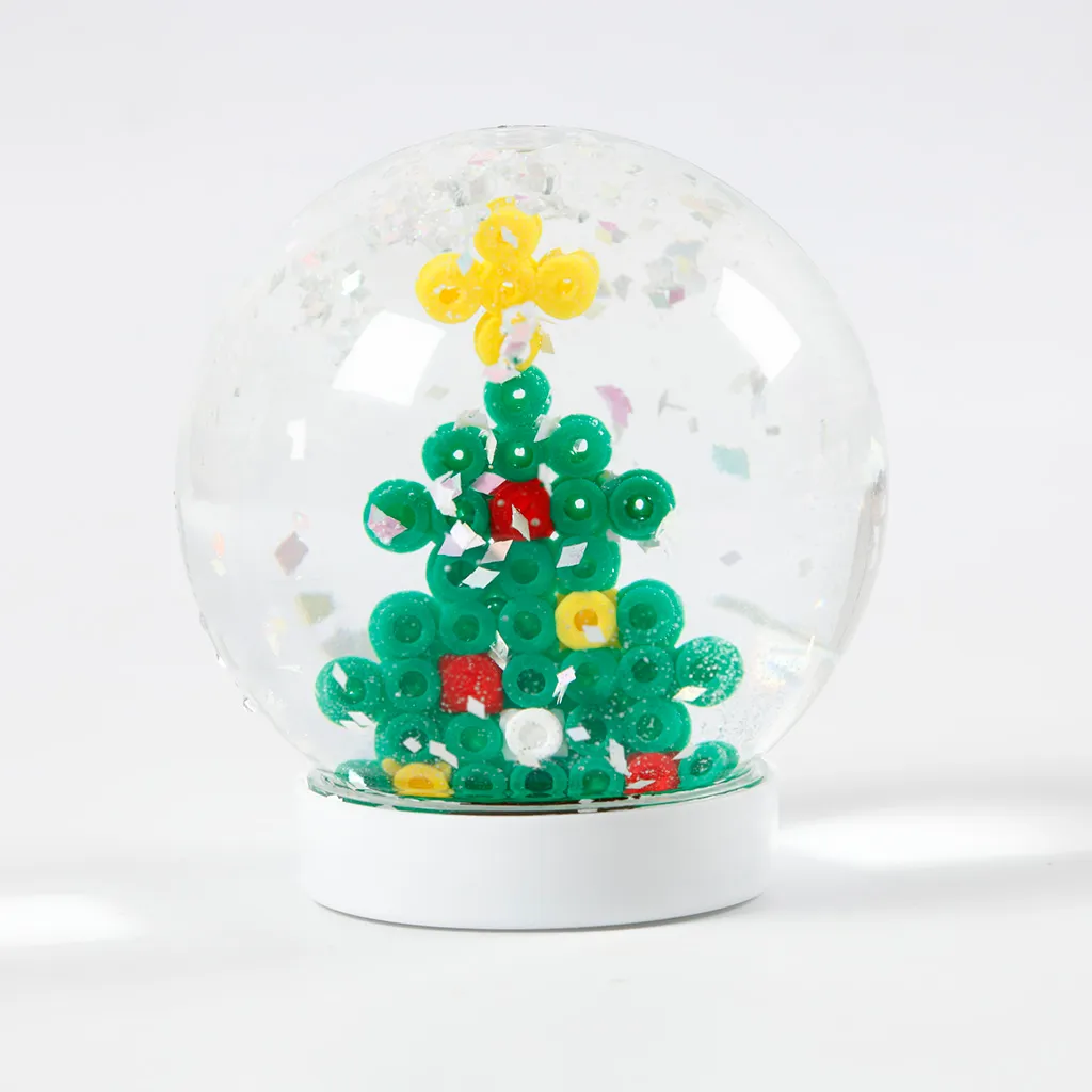 Een voltooide sneeuwbol op een witte voet met daarin een groene kerstboom van strijkkraaltjes, gedecoreerd met een gele ster en kleurrijke kerstballen, omringd door dwarrelende glitterstukjes.
