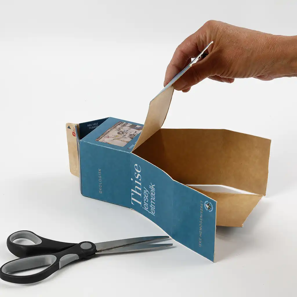 Een hand knipt met een schaar de boven- en zijkanten van een blauw melkpak open op een witte achtergrond.
