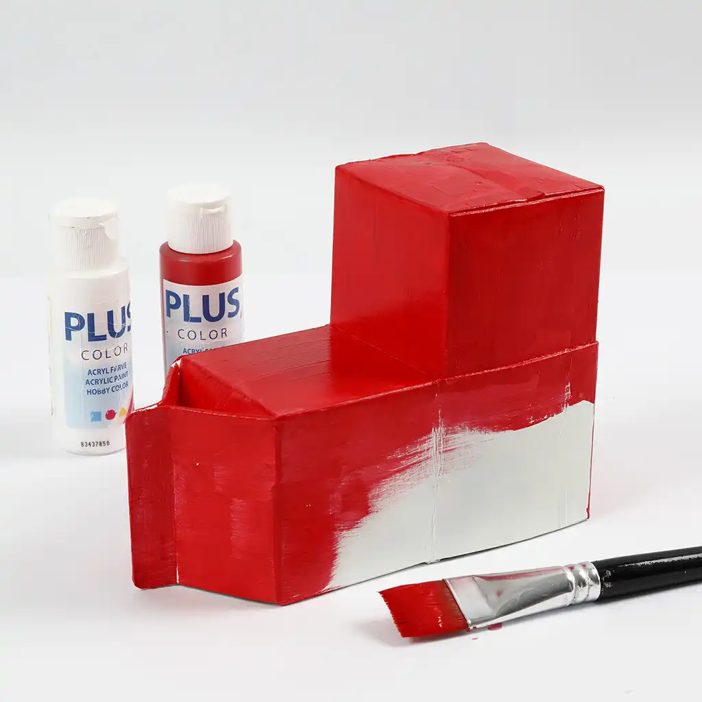 De basiselementen van de trein worden rood geschilderd met acrylverf en een brede kwast. Ernaast staan flesjes rode en witte Plus Color acrylverf.
