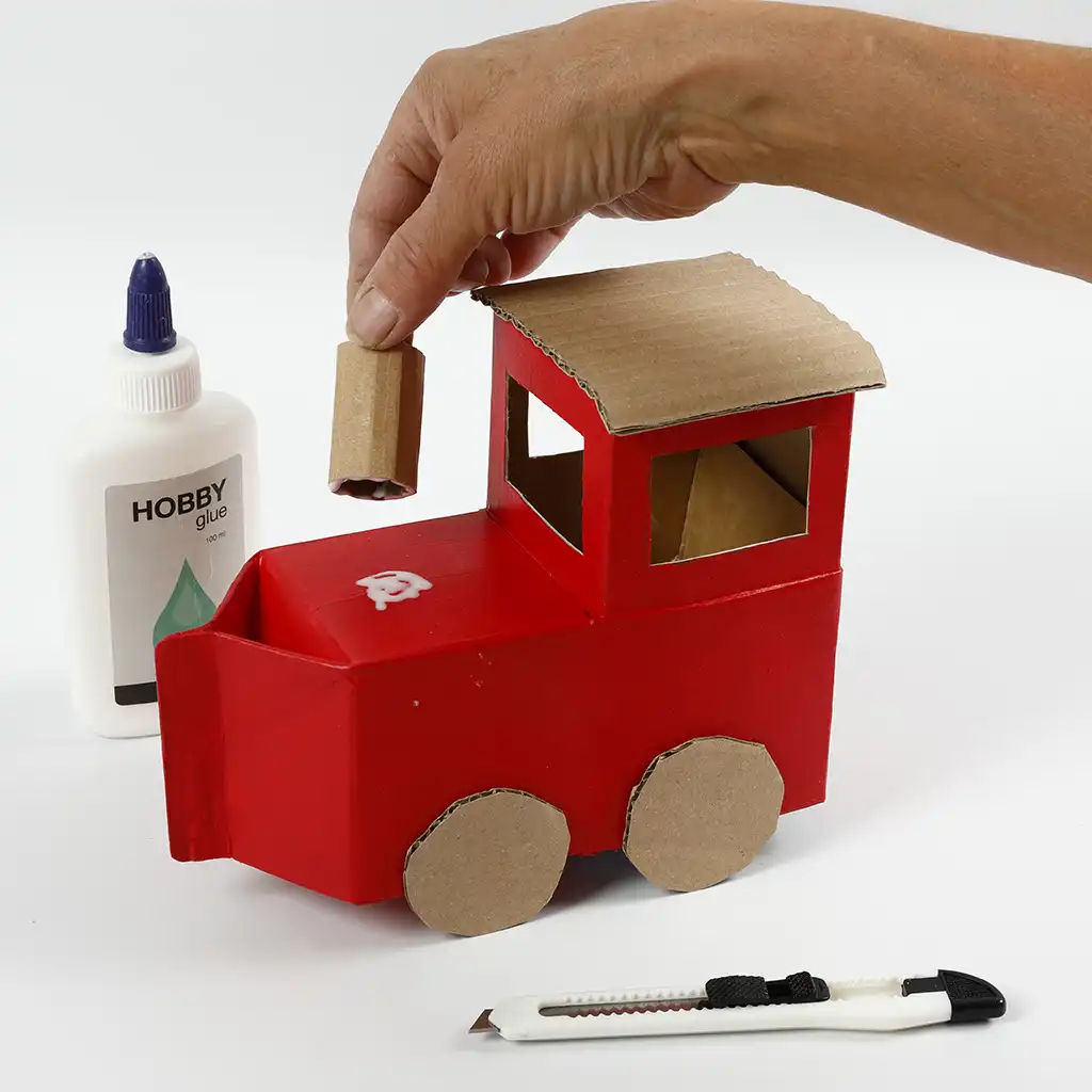 Een hand plaatst een opgerold stukje karton als pijp op de rode neus van de trein. De kartonnen wielen en het dakje zijn al vastgelijmd.