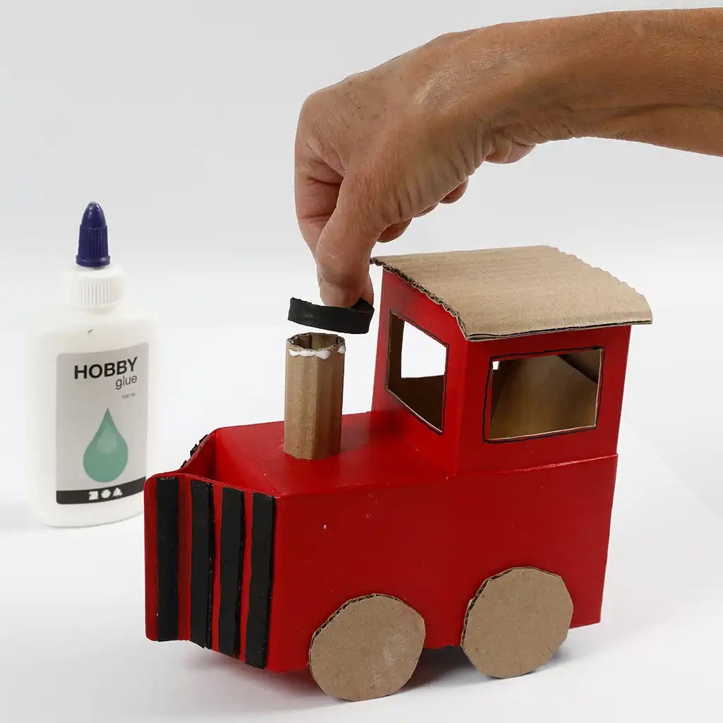Een hand plaatst een zwart geverfd kartonnen randje bovenop de stoompijp van de rode trein.
